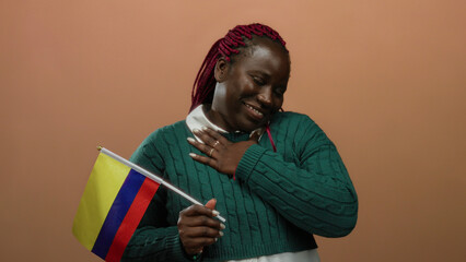 Fototapeta premium Woman holding colombian flag over isolated brown background, expressing solemn emotion, wearing green sweater, highlighting cultural pride and emotional connection.