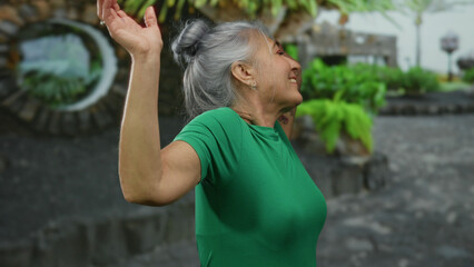 Senior woman with grey hair joyfully raising arms in a green shirt on a vibrant street dotted with lush greenery, showcasing her expressive personality and carefree spirit outdoors.
