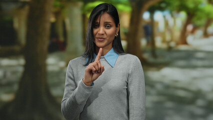 Young latin woman in a park gesturing with her finger while expressing negation, surrounded by trees and natural outdoor scenery, conveying emotion and contemplation.