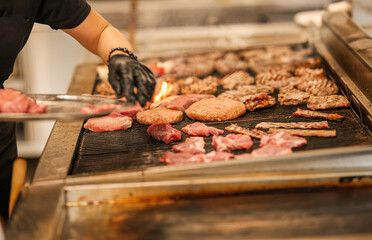 Chef placing raw meat onto a hot grill in a professional kitchen. A variety of meats are sizzling over open flame with smoke rising in a busy cooking environment.