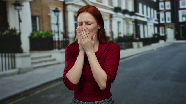 Woman sneezing on city street with red hair and red shirt, showing discomfort during outdoor allergy season.