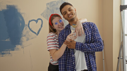 Couple painting new home together smiling and embracing while holding paintbrush in living room with heart painted on wall showcasing love and warmth indoors.