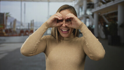 Woman outdoors making a playful binoculars gesture with her hands while smiling in an urban street...