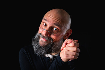 Portrait of a cunning-looking bald man with gray beard and glasses, clasping hands. Dark background. Mischievous or scheming expression. Concept of plotting, sarcasm, or ironic humor