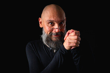 Portrait of a cunning-looking bald man with gray beard and glasses, clasping hands. Dark background. Mischievous or scheming expression. Concept of plotting, sarcasm, or ironic humor