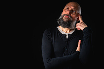 Thoughtful bald man with beard and glasses looking up, touching temple, deep in thought, isolated on black background. Concept of ideas, planning, decision-making