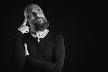 Black and white portrait, pensive bald man with beard and glasses looking up, touching temple, deep in thought, isolated on black background. Concept of ideas, planning, decision making