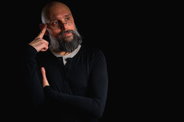 Thoughtful bald man with beard and glasses looking up, touching temple, deep in thought, isolated on black background. Concept of ideas, planning, decision-making