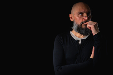 Thoughtful bald man with beard and glasses looking up, touching temple, deep in thought, isolated on black background. Concept of ideas, planning, decision-making