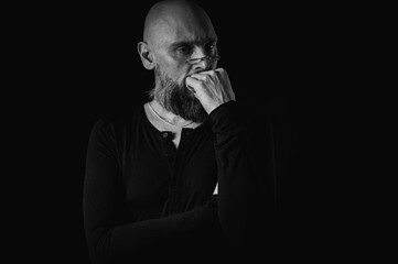 Black and white portrait, pensive bald man with beard and glasses looking up, touching temple, deep in thought, isolated on black background. Concept of ideas, planning, decision making
