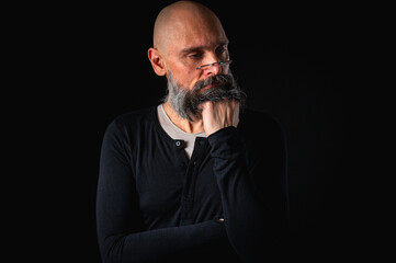 Thoughtful bald man with beard and glasses looking up, touching temple, deep in thought, isolated on black background. Concept of ideas, planning, decision-making