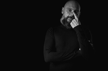 Black and white portrait, pensive bald man with beard and glasses looking up, touching temple, deep in thought, isolated on black background. Concept of ideas, planning, decision making