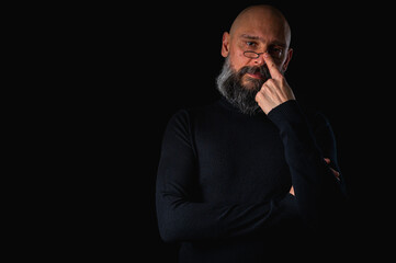 Thoughtful bald man with beard and glasses looking up, touching temple, deep in thought, isolated on black background. Concept of ideas, planning, decision-making