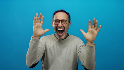 Mature hispanic man expressing joy and excitement with raised hands against a vibrant blue...