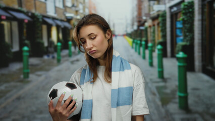 Young woman holding a soccer ball on a city street appears thoughtful wearing a striped scarf in an...
