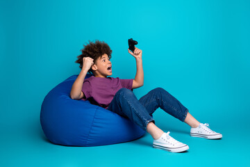 Joyful young boy celebrating a gaming victory while sitting on a blue beanbag in a stylish and...