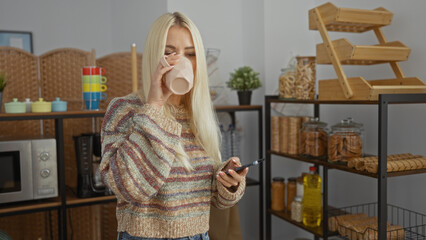 Young woman drinking coffee while holding smartphone in a cozy home kitchen, surrounded by various jars and colorful decor elements creating a relaxed and inviting atmosphere.