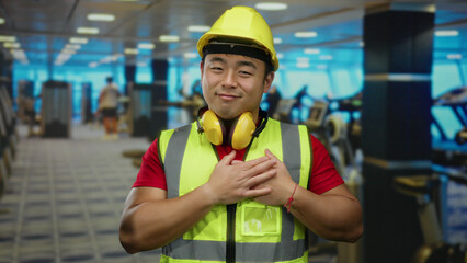 Man in gym wearing hard hat and reflective vest with headphones smiling and crossing hands on chest, expressing gratitude amid exercise equipment in bright indoor setting.