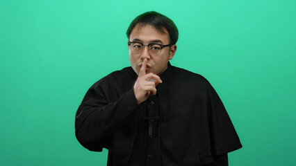 Asian priest gesturing silence against a bright green background, wearing glasses and a clerical...