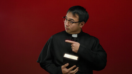 Asian priest in black robe holding bible and pointing on red background, exuding confidence and faith.