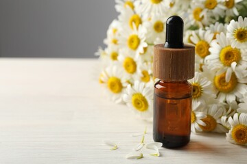 Bottle of essential oil and chamomile flowers on light wooden table, closeup. Space for text
