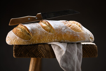Crusty sourdough baguette bread with cracked crust and rugged texture on a rustic wooden stool with knife and linen towel. Country-style fermented dough framed for artisanal food stock