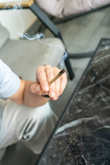 A man holds a burning cannabis joint between his fingers. Smoke rises in soft curls, with a marble table and relaxed interior suggesting a peaceful indoor setting.