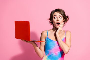 Charming young woman in vibrant summer outfit holding red laptop with surprised expression on pink...