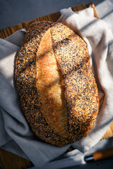 Crispy loaf of sourdough bread with cracked crust and rugged texture, covered with different grains and wrapped in a linen towel close up