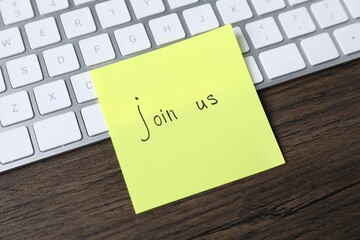Paper note with phrase Join Us and keyboard on wooden table, closeup