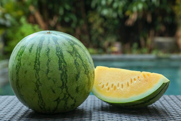 Whole and cut yellow watermelons on wicker table near swimming pool outdoors, closeup