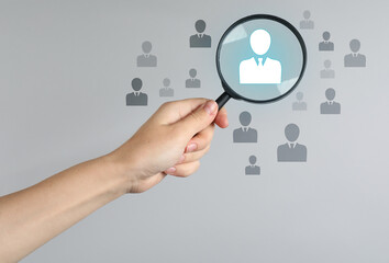 Employee search. HR manager holding magnifying glass over human icons against grey background, closeup