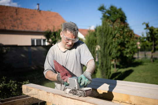 Carpenter sanding wooden plank outdoors using belt sander wearing safety glasses