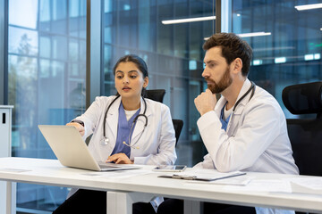 Two doctors, one female and one male, are looking at a laptop screen in a modern office setting,...