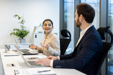 An enthusiastic businesswoman gestures while discussing a project with her colleague in a modern office environment, using laptops.