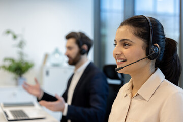 A smiling woman wearing a headset works at a call center alongside a colleague, both engaged in a...