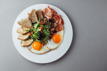 Healthy breakfast with eggs, bacon, grilled chicken, beans, salad and bread on white plate – top view gourmet morning meal on gray background
