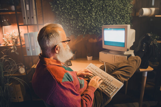 Elderly man with a beard coding on a vintage computer in a cozy evening home office setting