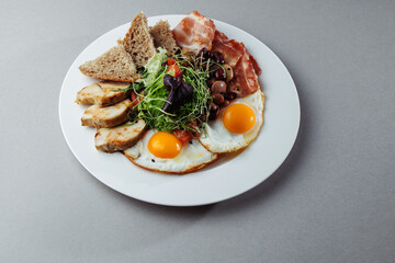 Healthy breakfast with eggs, bacon, grilled chicken, beans, salad and bread on white plate – top view gourmet morning meal on gray background