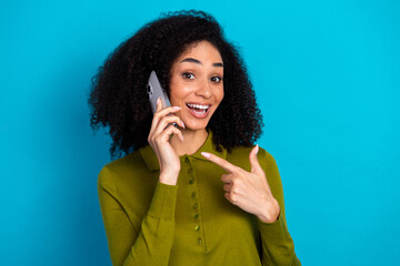 Happy young woman in casual khaki shirt joyfully pointing at smartphone against blue background