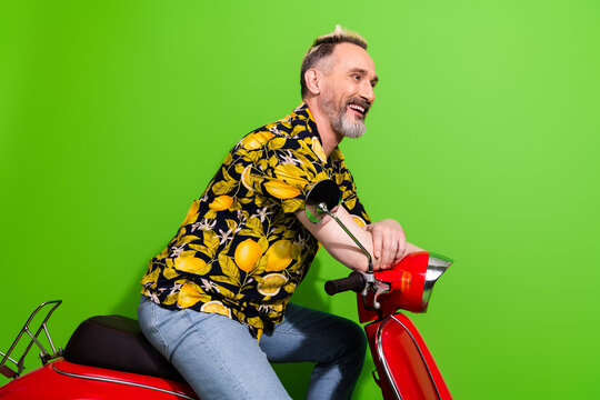 Mature man with stylish beard poses on a vibrant red scooter wearing a tropical print shirt, set against a lush green background