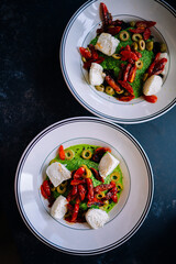 Two bowls of burrata with pesto, grilled red peppers, and green olives. Creative overhead flat lay food styling on dark background