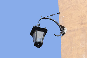 Old vintage lantern on wall. Classic black metal lamp on historical building. Bright sunny day and blue sky background. Retro city street architecture element. Outdoor lighting in urban space.