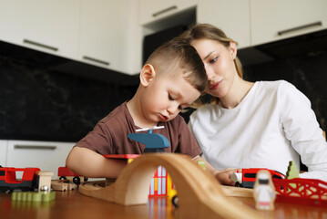 Young mother and her little son deeply focused while playing with a wooden toy train set at the kitchen table, sharing a quiet and thoughtful moment together.