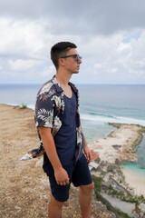 A young man in sunglasses and a tropical shirt stands on a cliff overlooking the ocean and Melasti beach on a cloudy day in Bali, Indonesia