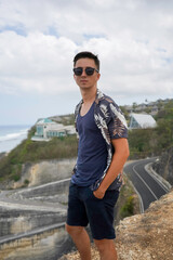 A young man in sunglasses and a tropical shirt stands on a cliff overlooking the ocean and Melasti beach on a cloudy day in Bali, Indonesia