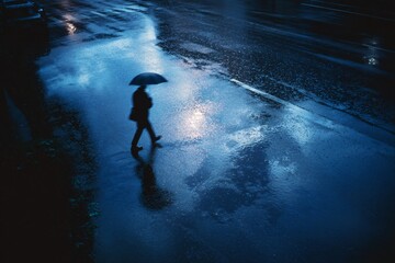 Obraz premium Solitary Individual Walks Under Umbrella on Rainy Night in Blue-Tinted Street