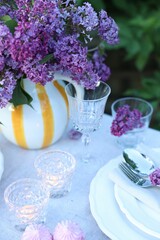 Beautiful lilac flowers, plates, cutlery, glasses and burning candles on table in garden, closeup