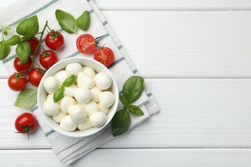 Tasty mozzarella cheese balls in bowl, basil and tomatoes on white wooden table, flat lay. Space for text