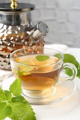 Aromatic lemon balm tea in glass cup, teapot and fresh leaves on white table, closeup